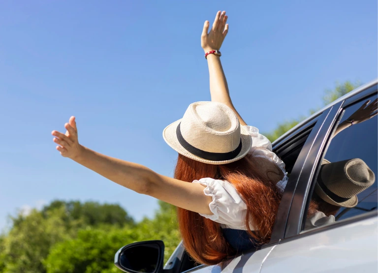 woman wearing hat, leaning out of car window with arms up in the air feeling breeze